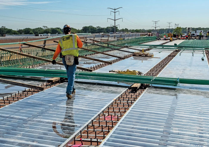 Construction workers install large steel beams on a highway bridge under clear skies, with vehicles in the background.