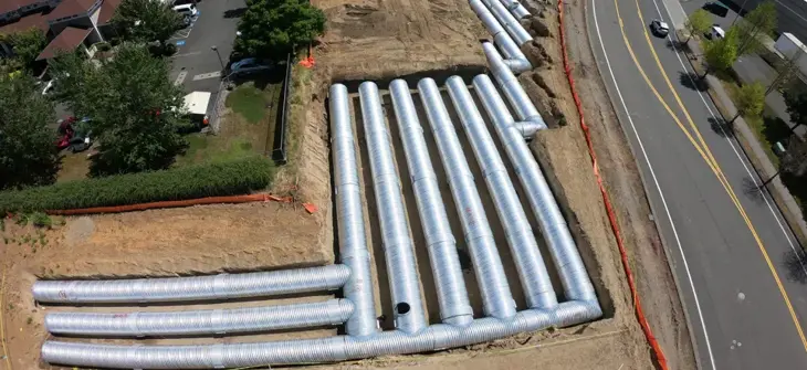 A construction site with large metal pipes laid out in parallel rows next to a road and nearby buildings.