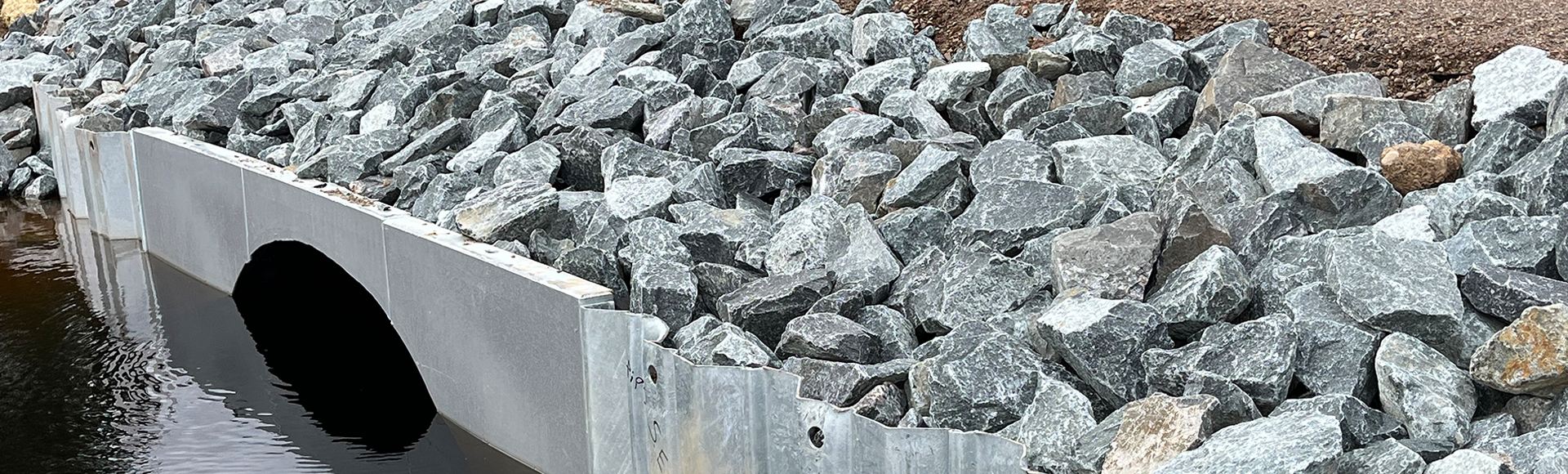 Culvert under a gravel road with large rocks lining the embankment, leading to water below.